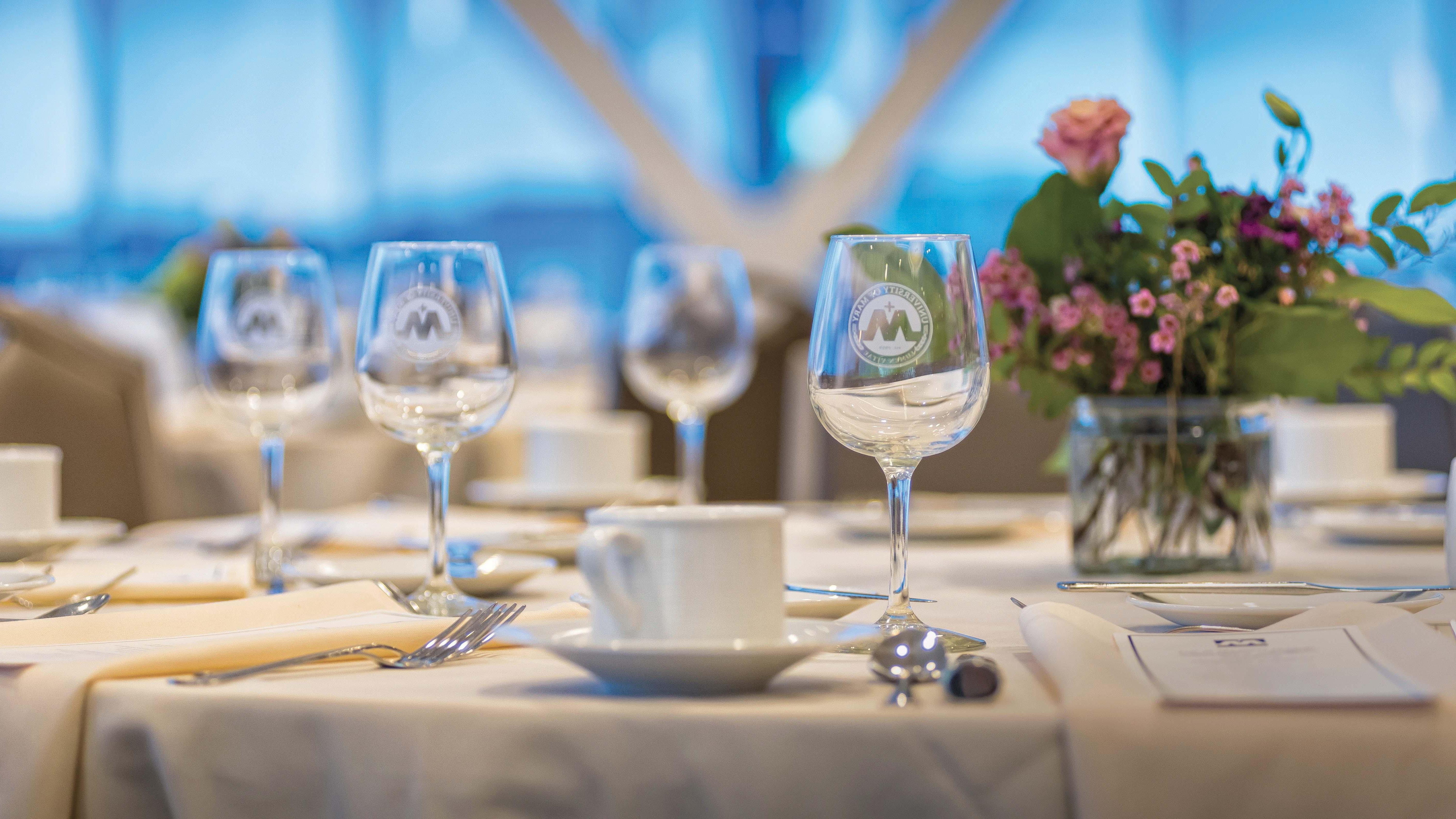 University of Mary wine glasses etched with its seal are set on a table with a centerpiece, menus, silverware, and napkins adding elegance to the formal dining decor inside Founders Hall on campus.